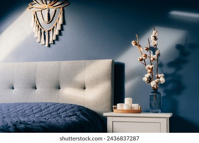 Stylish modern bedroom in dark colors. Cozy interior with navy blue walls, home decor. Bed with gray fabric headboard, blue blanket, bedside table, vase with natural cotton flowers, candles on tray