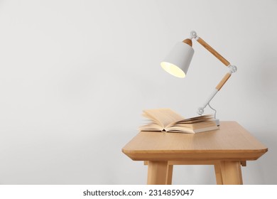 Stylish modern desk lamp and open book on wooden table near white wall, space for text