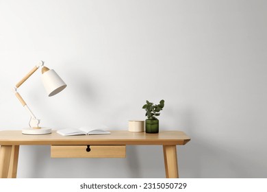 Stylish modern desk lamp, open book and plant on wooden table near white wall, space for text