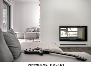 Stylish modern interior with light walls and a parquet. There are gray sofas with pillows and a guitar, fireplace, table, large windows. Horizontal.