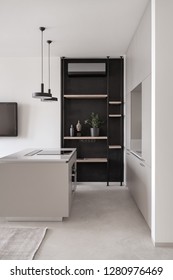 Stylish Modern Kitchen With Light Walls And A Gray Floor With A Carpet. There Is A Kitchen Island With A Cooking Surface, Lockers, Shelves, Plant, Hanging Black Lamps, Dark TV, Conditioner. Vertical.