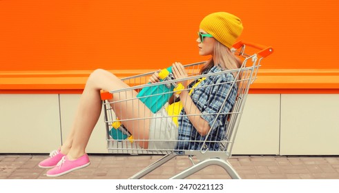 Stylish modern young woman sitting in shopping trolley cart with skateboard in colorful clothes on orange background on city street - Powered by Shutterstock