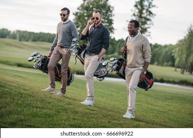 stylish multiethnic friends holding bags with golf clubs and walking on golf course  - Powered by Shutterstock