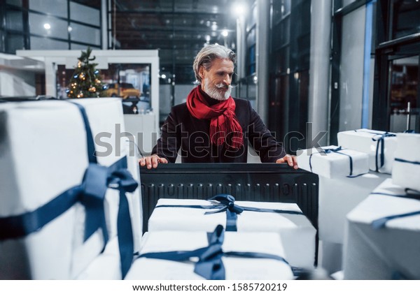 Stylish old man\
with grey hair and mustache packing presents in gift boxes into\
car. Christmas tree at\
background.