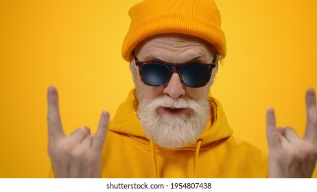 Stylish Old Man Showing Rock Gesture Indoors. Cheerful Elderly Guy Posing For Camera On Yellow Studio Background. Joyful Senior Male Person In Sunglasses Spending Time Inside.