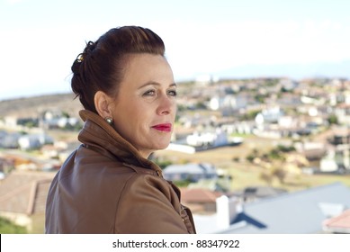 Stylish Older Caucasian Woman In A Brown Leather Jacket, Posing Casually Outdoors, Overlooking Some Houses