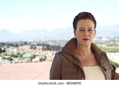 Stylish Older Caucasian Woman In A Brown Leather Jacket, Posing Casually Outdoors, Overlooking Some Houses