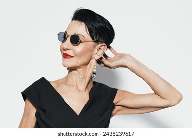 Stylish older woman wearing sunglasses and black dress posing for camera in front of white wall - Powered by Shutterstock