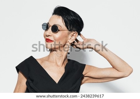 Stylish older woman wearing sunglasses and black dress posing for camera in front of white wall