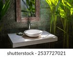 Stylish outdoor bathroom with a stone sink, rustic mirror, and lush tropical strelitzia reginae plants in natural sunlight. Jungle village interior design