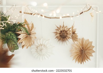 Stylish paper christmas stars and lights hanging on white wall background. Scandinavian festive decoration in boho room. Handmade paper swedish stars and garland. Atmospheric christmas time - Powered by Shutterstock