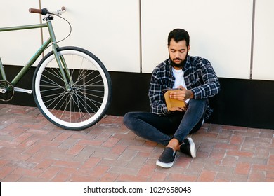 Stylish Pensive Bearded Man Sitting With Computer Tablet Near The Bike On The Ground, Reading Something, Wearing Trendy Clothes. Outside.