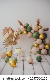 Stylish Photo Zone With A Cake And An Arch Of Beige, Green And Gold Balloons. Light Background White Wood Floor.