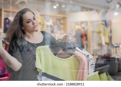 Stylish Plus Size Female Shopaholic Choosing Fashion Clothes At Store Boutique. Focused Blonde Woman Buyer Enjoying Shopping, Purchasing Goods Trendy Clothes At Modern Center Mall During Discount