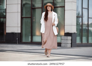 Stylish Plus size woman walks confidently outdoors in urban city, wearing beige dress, white blazer, wide brimmed hat, woven handbag. Model with long brown hair worn loose, calm, composed expression - Powered by Shutterstock