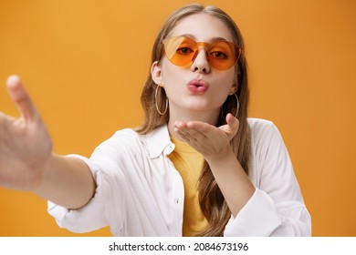 Stylish Popular Girl Sending Air Kiss At Camera Taking Selfie Or Recording Vlog Working As Freelance Travel Advisor Taking Pictures Against Orange Background Tender And Feminine Pulling Hand Forward