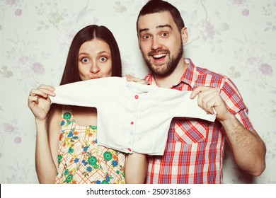 Stylish Pregnancy Concept: Portrait Of Funny Couple Of Hipsters (husband And Wife) In Trendy Clothes (shirt, Dress, Jeans) Holding Small Size Baby Shirt. Vintage (retro) Style. Studio Shot