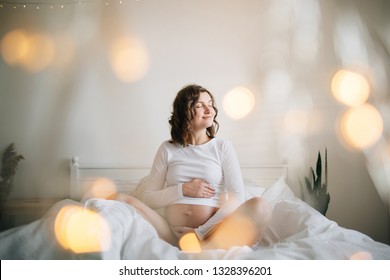 Stylish Pregnant Mom Hugging Belly With Love And Care, Waiting For Baby. Motherhood Concept, Magic Time. Happy Pregnant Woman In White Holding Belly Bump And Relaxing On White Bed At Home