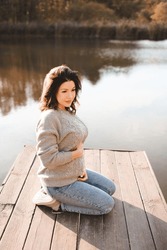 Stylish Pregnant Woman Wearing Knitted Sweater And Pants Sitting On Wooden Pier Over Lake Nature Background. Motherhood. Maternity. 