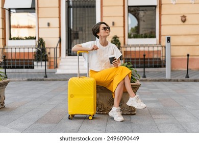 stylish pretty woman traveling with yellow suitcase in Europe in trend summer outfit street style, smiling happy sunny, skirt, dress and top, sitting waiting drinking coffee, wearing sunglasses - Powered by Shutterstock