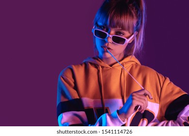 Stylish pretty young 20s fashion teen girl model wear glasses chew bubble gum look at camera stand at purple studio background, igen teenager with bubblegum in trendy night 80s party light portrait
