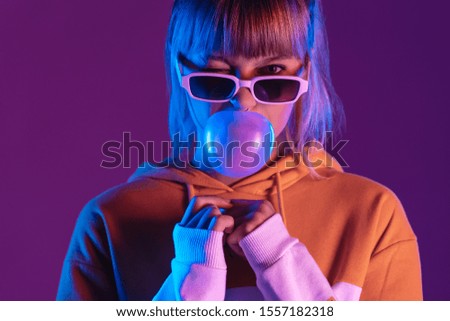 Stylish pretty young 20s fashion teen girl model wear glasses blowing bubble gum winking looking at camera stand at purple studio background, igen teenager in trendy light 80s 90s concept, portrait