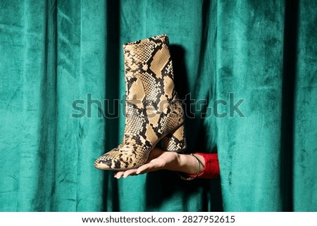 Stylish python print ankle boot held by a hand in a red sleeve against a luxurious green velvet curtain