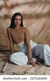 Stylish Queer Person In White Pants Sitting On Abstract Brown Background