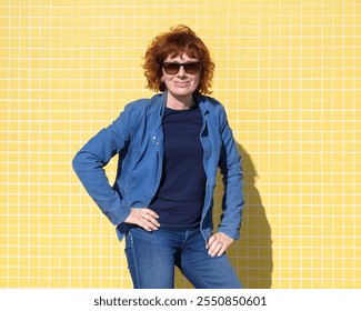 Stylish redhead woman in sunglasses and denim jacket posing confidently against a vibrant yellow tiled wall - Powered by Shutterstock