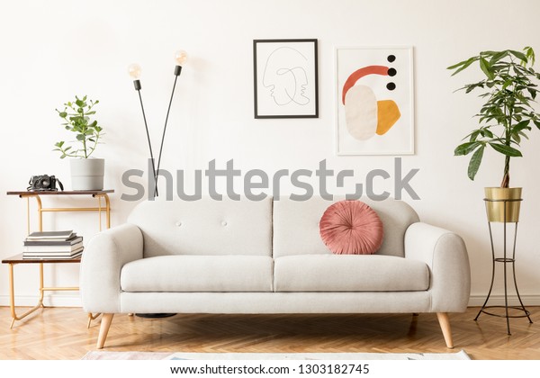 Stylish
retro and vintage interior of sitting room with design  sofa, gold
bookstand and mock up posters frames on the white walls. Interior
design with brown wooden parquet and
plants.