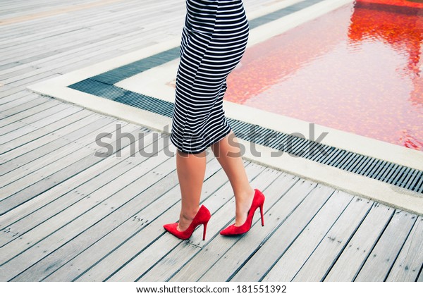 Stylish retro woman outfit with black and white\
striped dress red high heels classic pumps, posing near creative\
red pool.