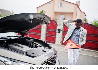 Stylish and rich african american man stand in front of a broken suv car needs assistance looking under opened hood and hold e,ergency triangle.