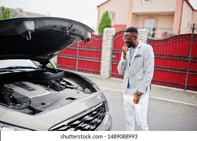 Stylish and rich african american man stand in front of a broken suv car needs assistance looking under opened hood.