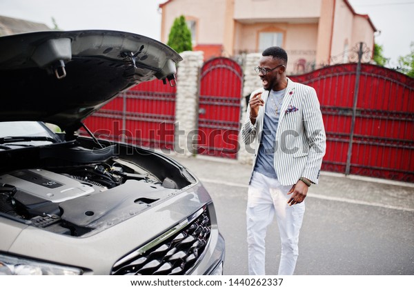 Stylish and\
rich african american man stand in front of a broken suv car needs\
assistance looking under opened\
hood.