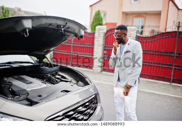 Stylish and\
rich african american man stand in front of a broken suv car needs\
assistance looking under opened\
hood.