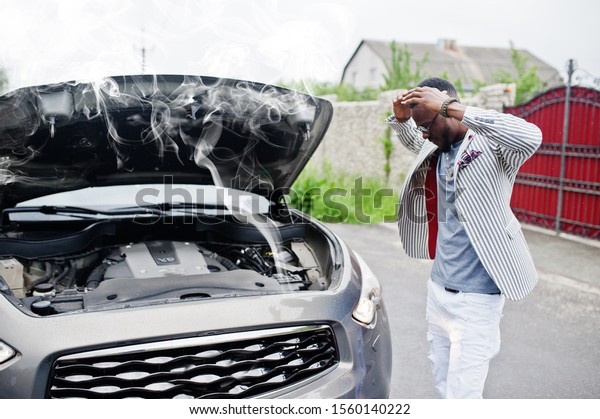 Stylish and rich african american man stand in\
front of a broken suv car needs assistance looking under opened\
hood with smoke.