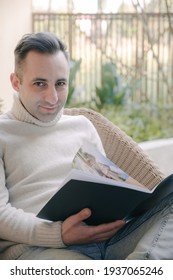 Stylish Rich Caucasian Man Sits On Summer Terrace Near Pool And Reads Real Estate Magazine To Choose Property To Buy. Businessman Spends Leisure Time Outside Reading Journal Outside His Mansion.