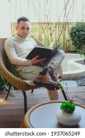 Stylish Rich Caucasian Man Sits On Summer Terrace Near Pool And Reads Real Estate Magazine To Choose Property To Buy. Businessman Spends Leisure Time Outside Reading Journal Outside His Mansion.