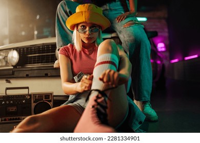 A stylish roller skate girl is sitting on the ground tying her skate lace and looking at the camera with her friend sitting behind her on a hood of a car.