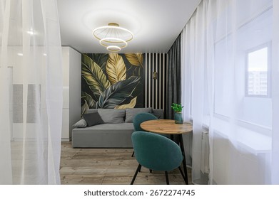 stylish room featuring sofa, table with two chairs, and striking leaf-themed wallpaper. space is bathed in light from window, softened by sheer curtains. A modern light fixture hangs from the ceiling