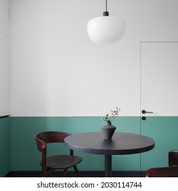 Stylish, round, wooden and dark dining table with decorative vase and modern chairs under new lamp in room with white and green wall
