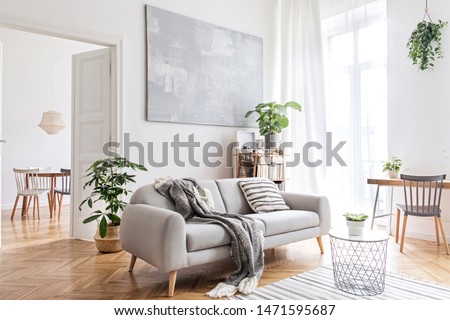 Stylish scandinavian living room with design furniture, plants, bamboo bookstand and wooden desk. Brown wooden parquet. Abstract painting on the white wall. Nice apartment. Modern decor of bright room