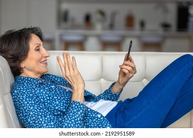 Stylish Senior Beautiful Woman With Mobile Phone Using Video Call To Chat With Friends While Lying On Sofa. Modern Older Woman Spending Weekend At Home, Relaxing Chatting With Friend Use Smartphone