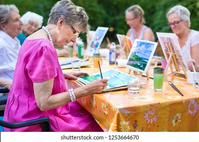 Stylish senior lady painting in art class with friends from her care home for the aged copying a painting with water colors on a canvas outdoors at a table in the garden. - Powered by Shutterstock