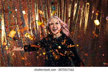 Stylish Senior Woman In Black Dress Throwing Confetti Against A Golden Backdrop