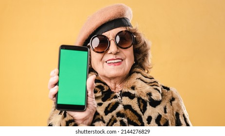 Stylish Senior Woman Showing A Mobile Phone With Green While Standing Over An Isolated Background.