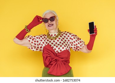 Stylish senior woman smiling and holding smartphone with blank screen on yellow background - Shutterstock ID 1682753074