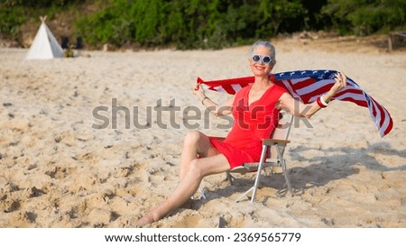 Stylish senior woman wearing united states flag and fashion sunglasses looking at the sea sky sitting on chair on beach. Vacation trip summer holiday.