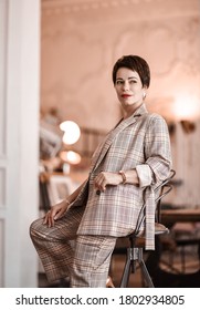 Stylish Short Haired Brunette Woman In Elegant Business Plaid Pantsuit With Rolled Up Sleeves Sits On A High Chair In Room Appartement Office Gallery. Business Elegance Concept