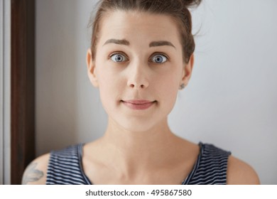 Stylish Shot Of Surprised Scared Caucasian Girl With Brown Hair, Charming Blue Eyes, White Skin. Young Female With Tattoo Looks Lovely In Her Natural Amusement With Closed Mouth, Raised Eyebrows.